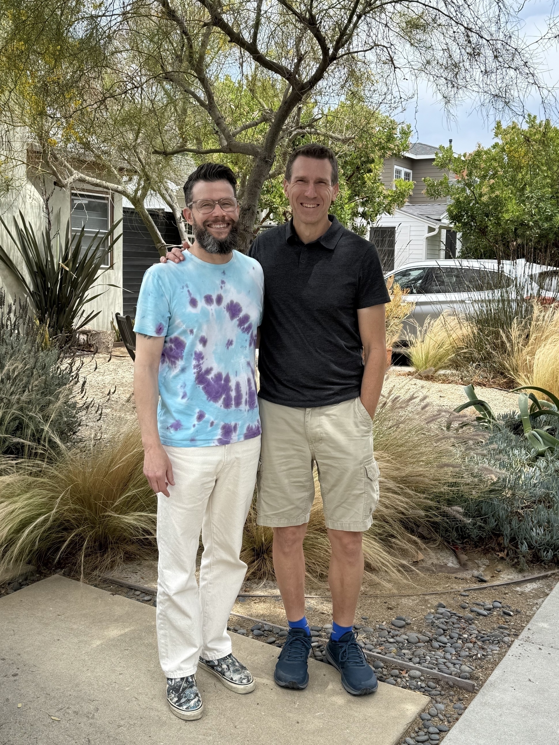 Auto-generated description: Two people stand smiling outdoors, surrounded by plants and trees, with one wearing a tie-dye shirt and the other in a black polo.