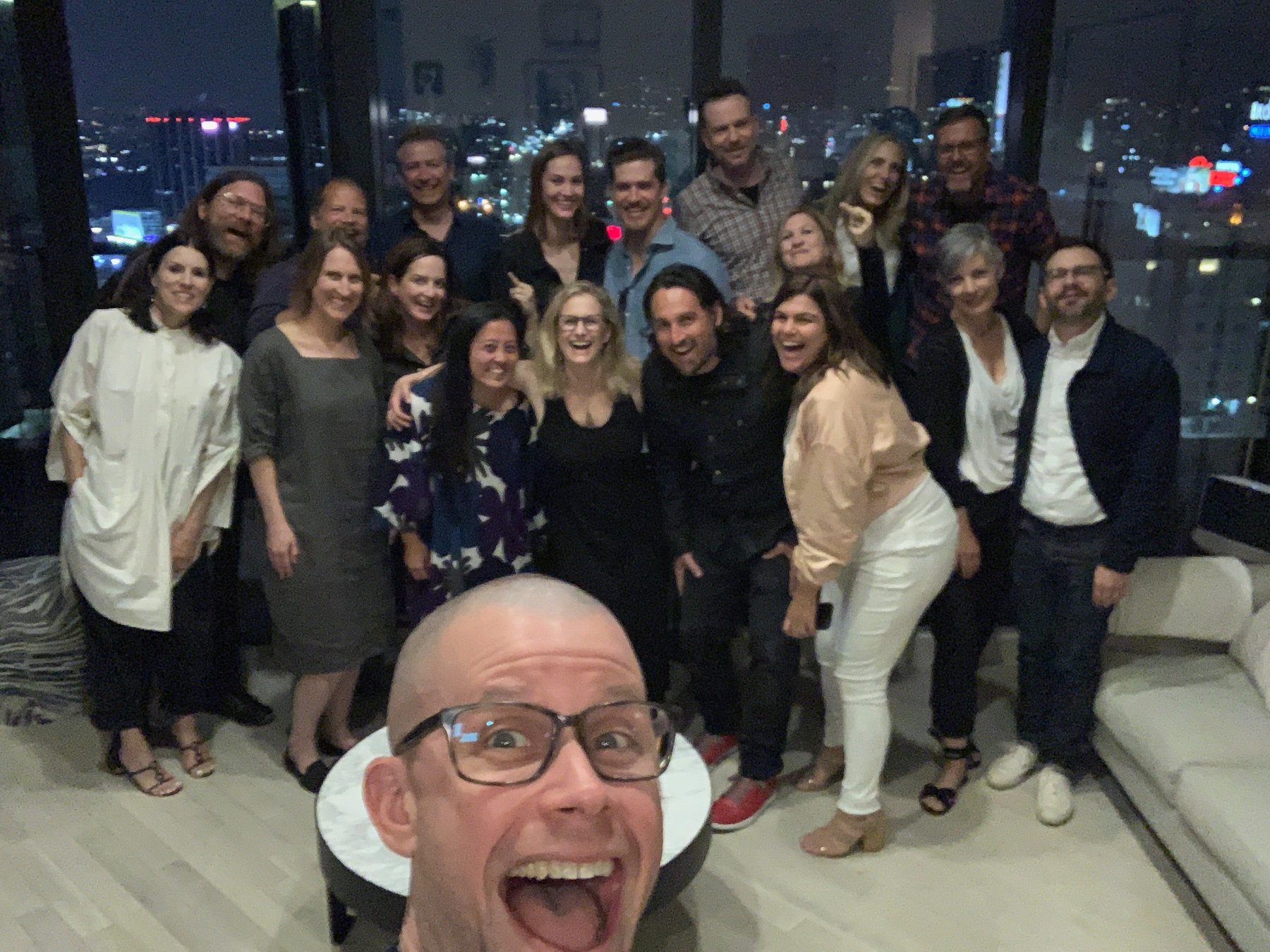 Auto-generated description: A group of people are posing together for a cheerful selfie at a nighttime gathering in a room with large windows overlooking a city.